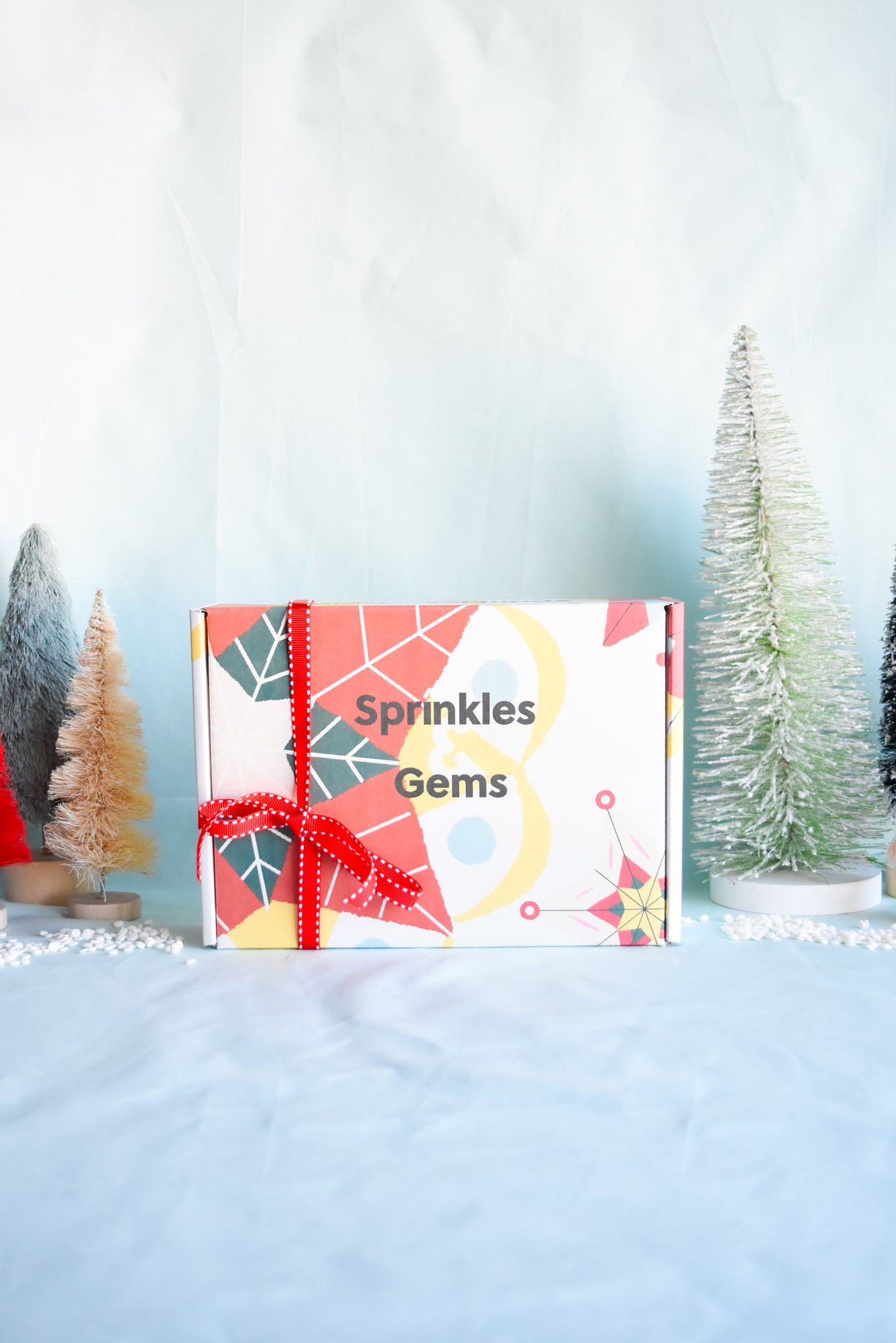 Sprinkles & Gems holiday-themed decorating kit box with colorful geometric design, wrapped with a red ribbon, surrounded by miniature Christmas trees. Perfect for festive cookie and cupcake decorating activities during the holiday season.