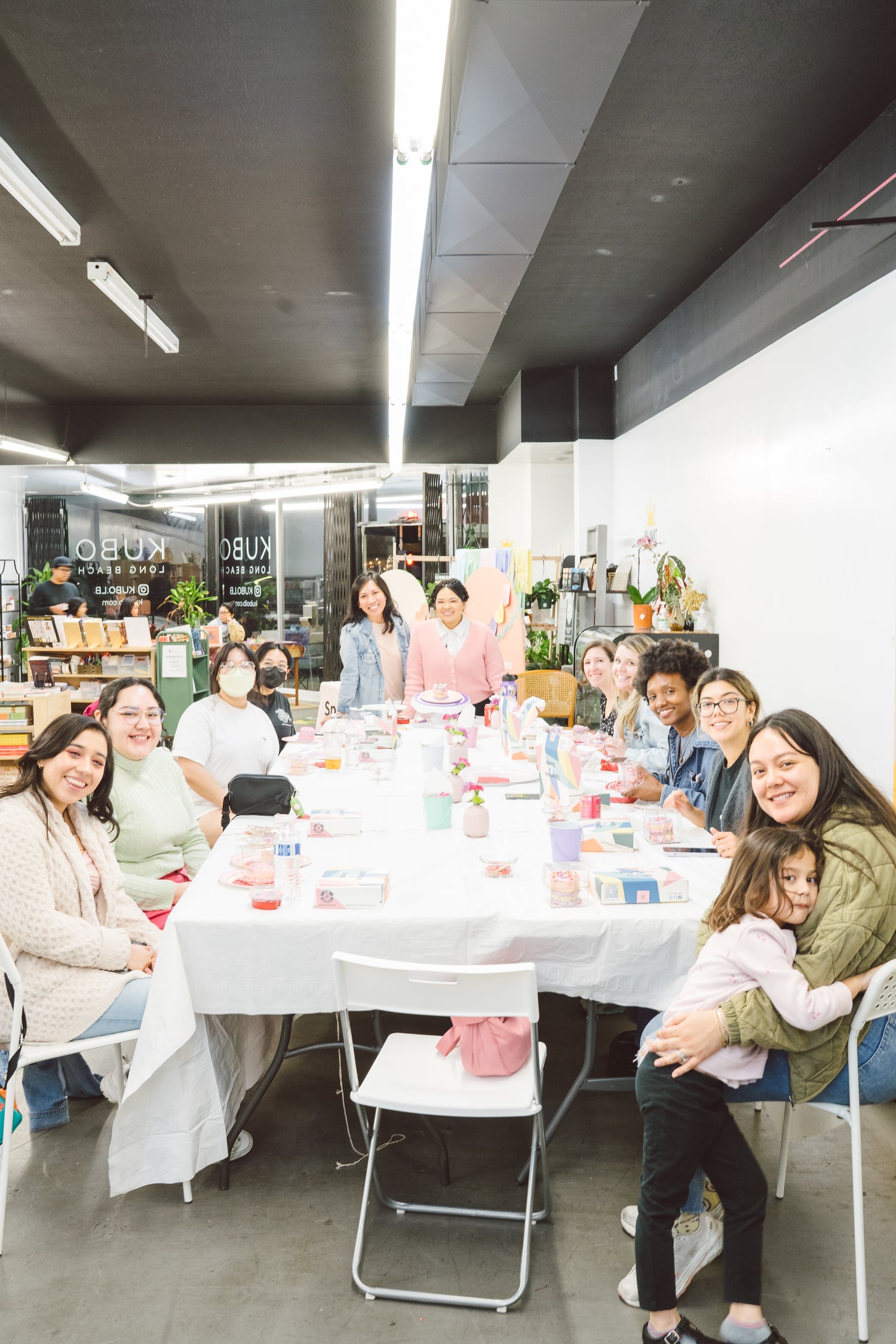 Group of women and children enjoying a fun decorating workshop hosted by Sprinkles & Gems at Kubo Long Beach. Participants are smiling around a table, working on their colorful mooncake decorating kits in a welcoming and creative environment.
